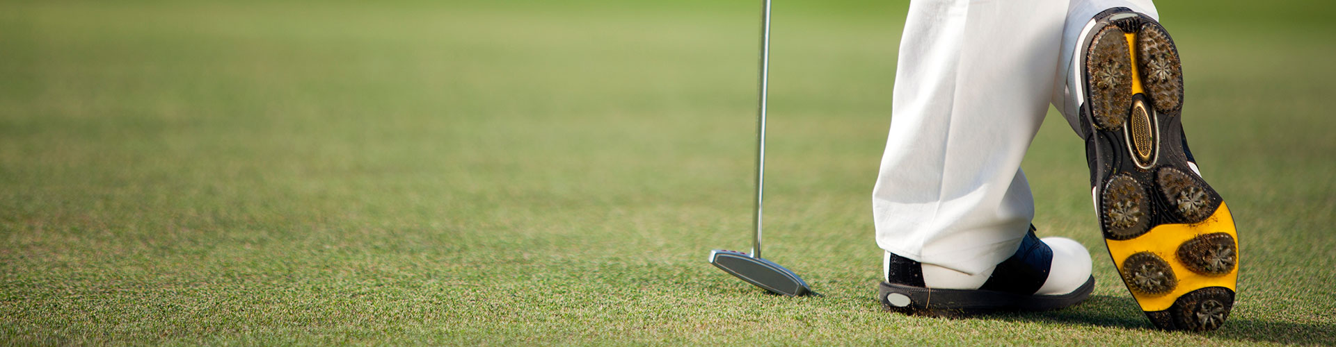 Golfer standing with one cleat facing camera and putter resting on the green.