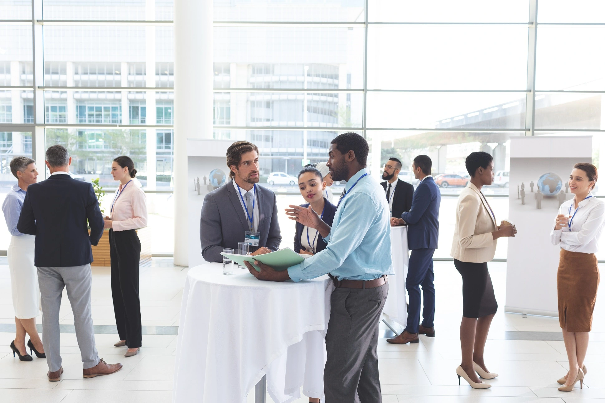A diverse group of people meeting in a room with large industrial windows and an urban background.