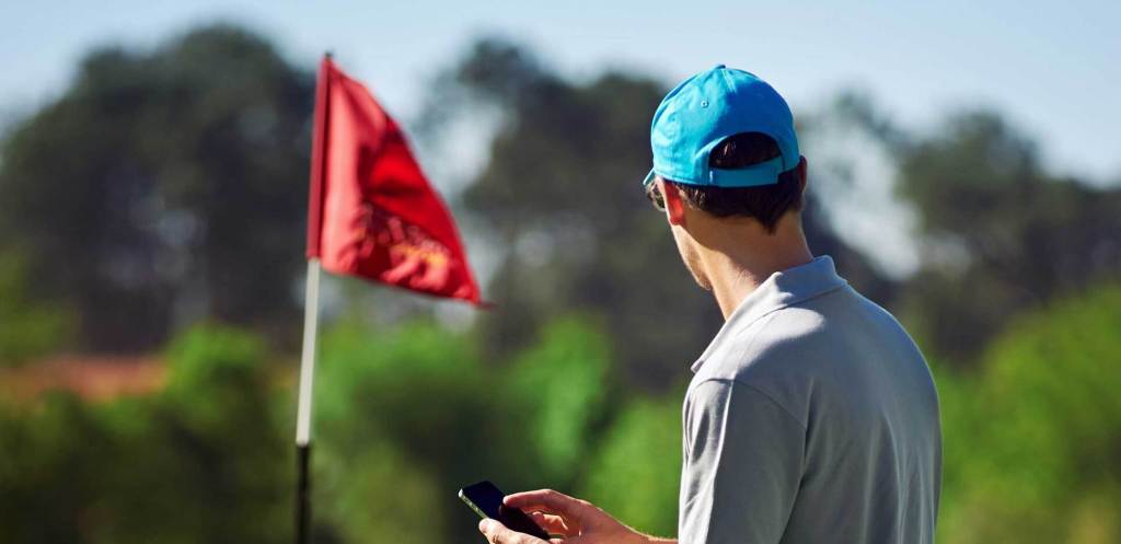 Golfer using phone on golf course with red flag over hole in distance 
